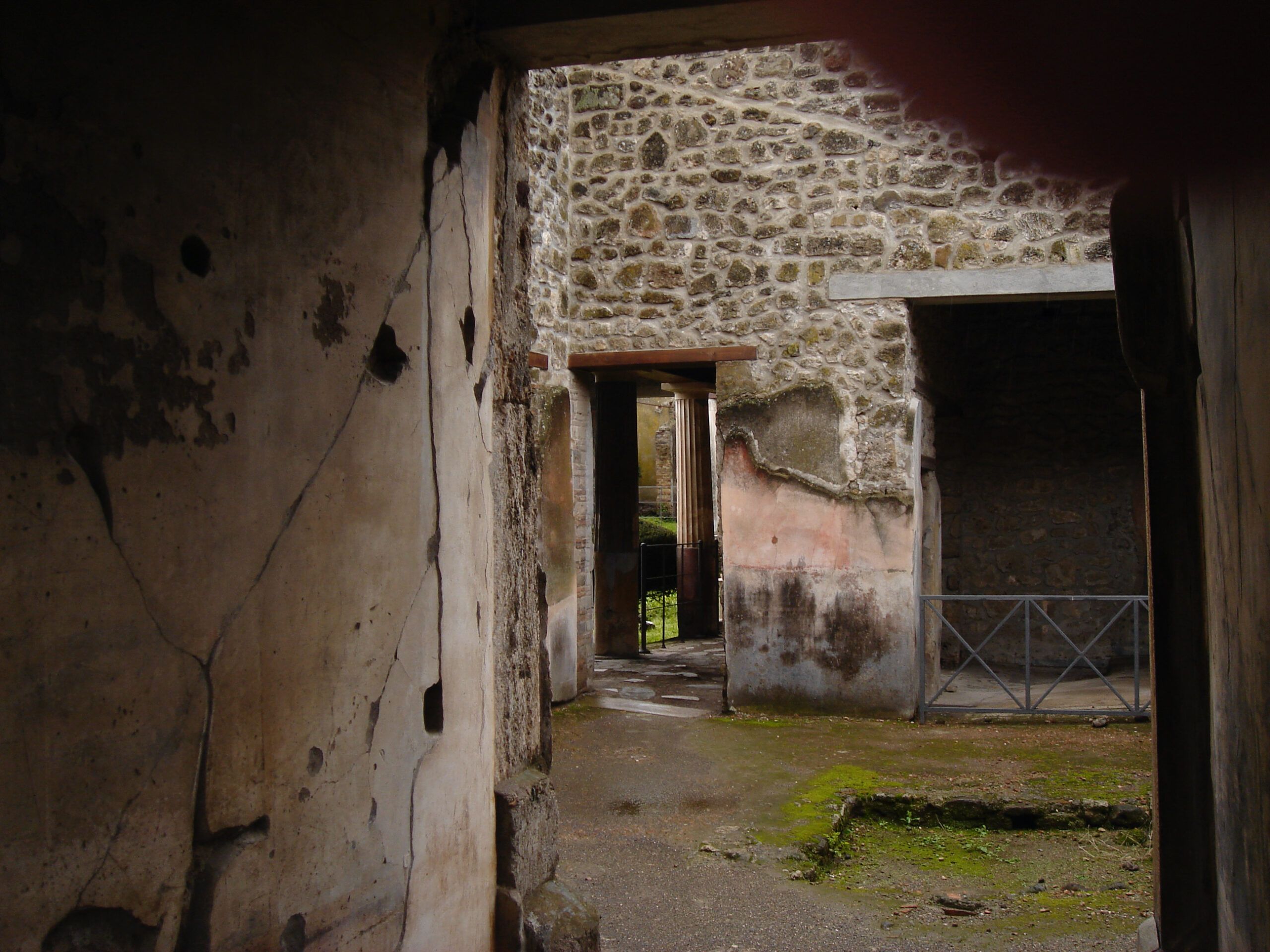 View through the entrance of a house in Pompeii, with cracked plaster walls and glimpses of inner rooms and a small garden.
