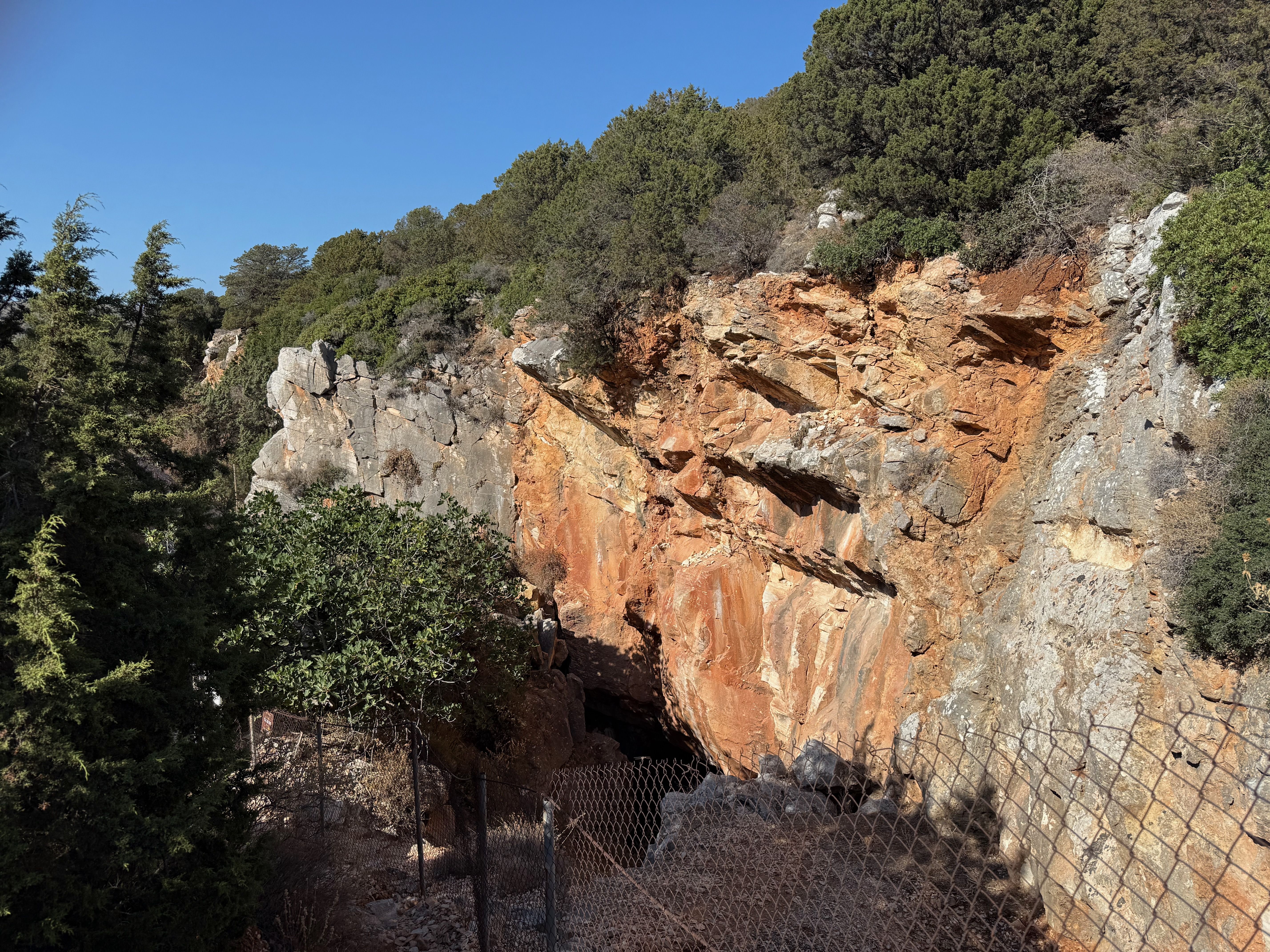A cliff wall, red with iron oxide, fenced off with a wire fence. A dark hole leads under the cliff wall into the quarry.