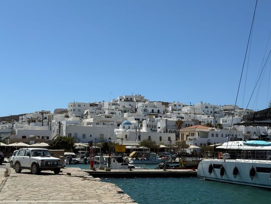 A pile of square white buildings set on a hill in the background; in the foreground a stone jetty with a yacht. There are some restaurant tables in behind the jetty.