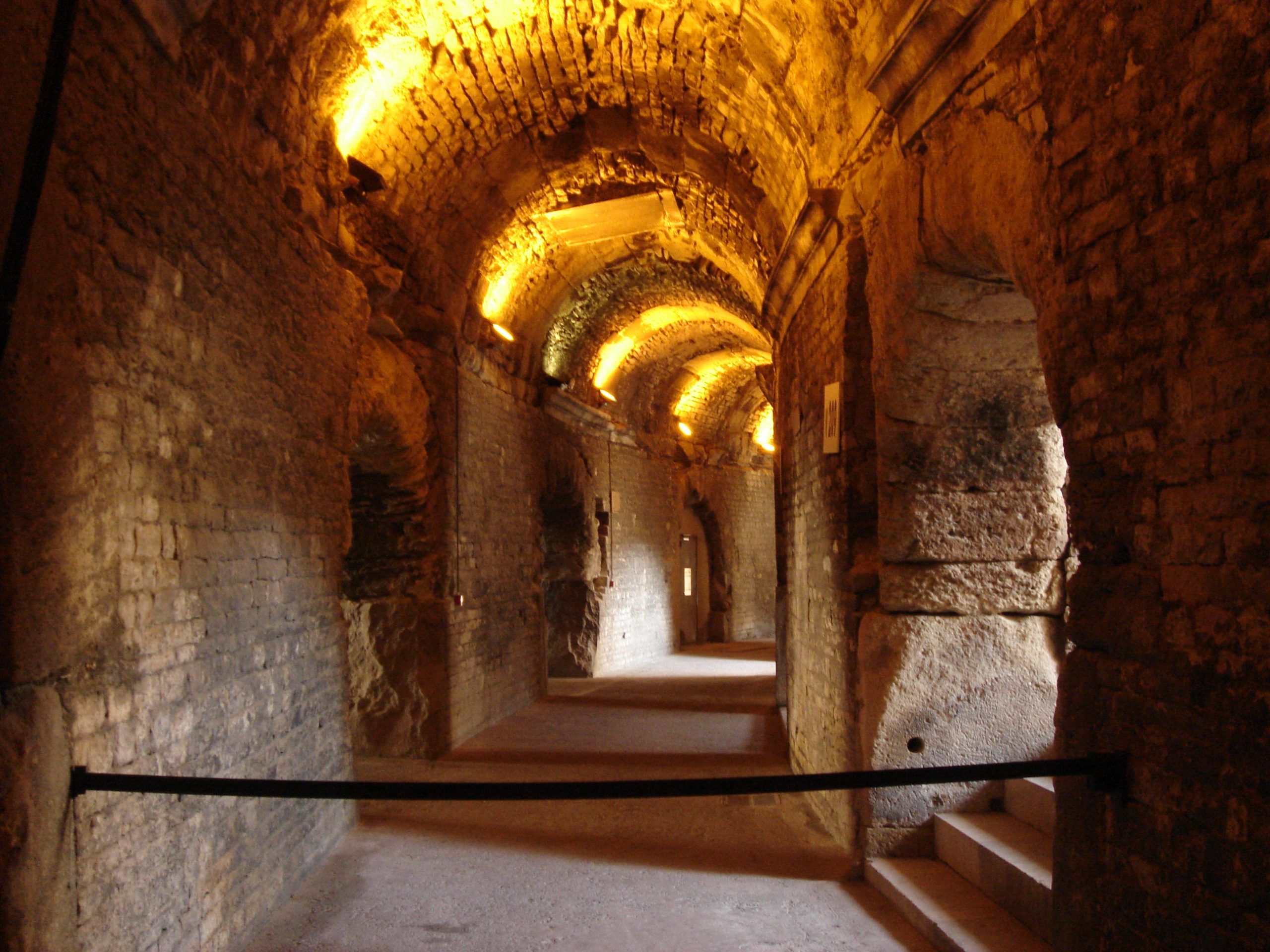 Interior corridor of the Roman amphitheatre in Nîmes, with vaulted stone ceiling lit by warm uplights.