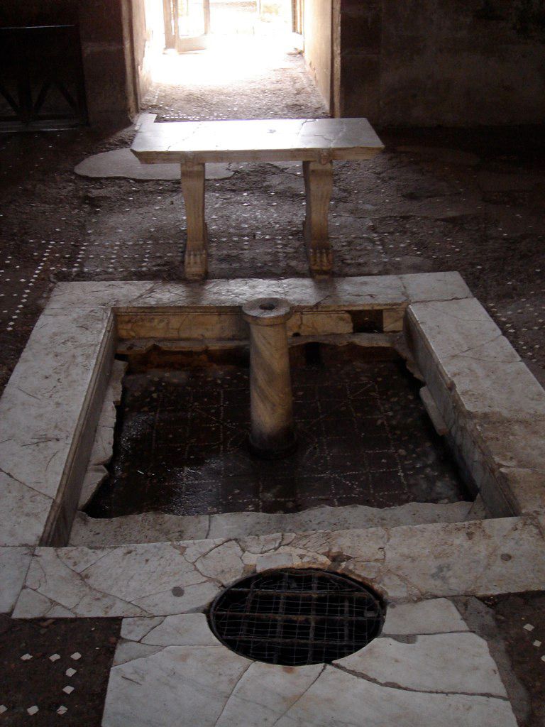 Marble-lined impluvium in a Roman house at Herculaneum, with a central column and a small table, illuminated by sunlight through the doorway.