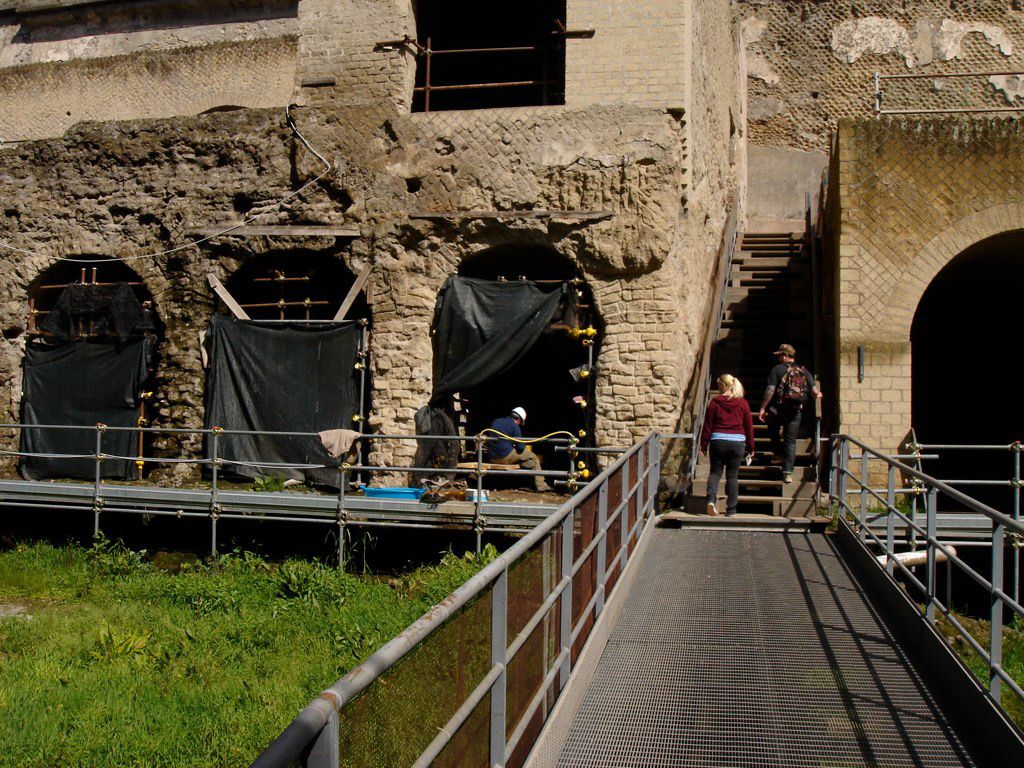Visitors ascending metal walkways beside an excavation site at Herculaneum, with partially covered archways and structural scaffolding.