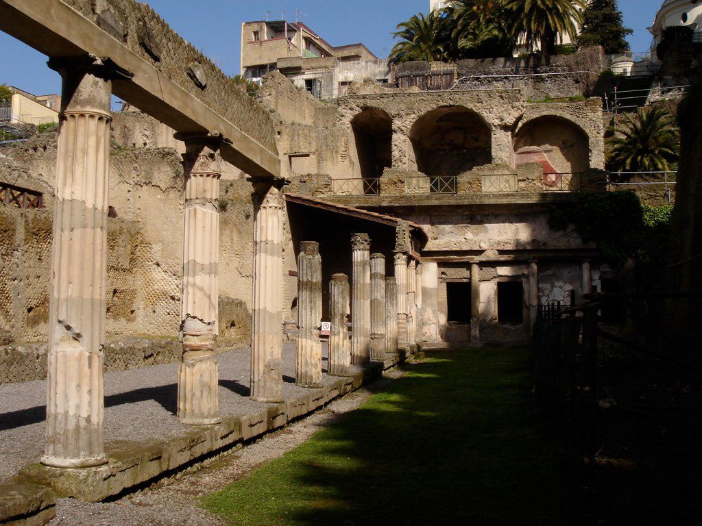 View of the upper terrace and colonnade of a grand Roman house in Herculaneum, framed by tall palm trees and multiple levels of arched rooms.