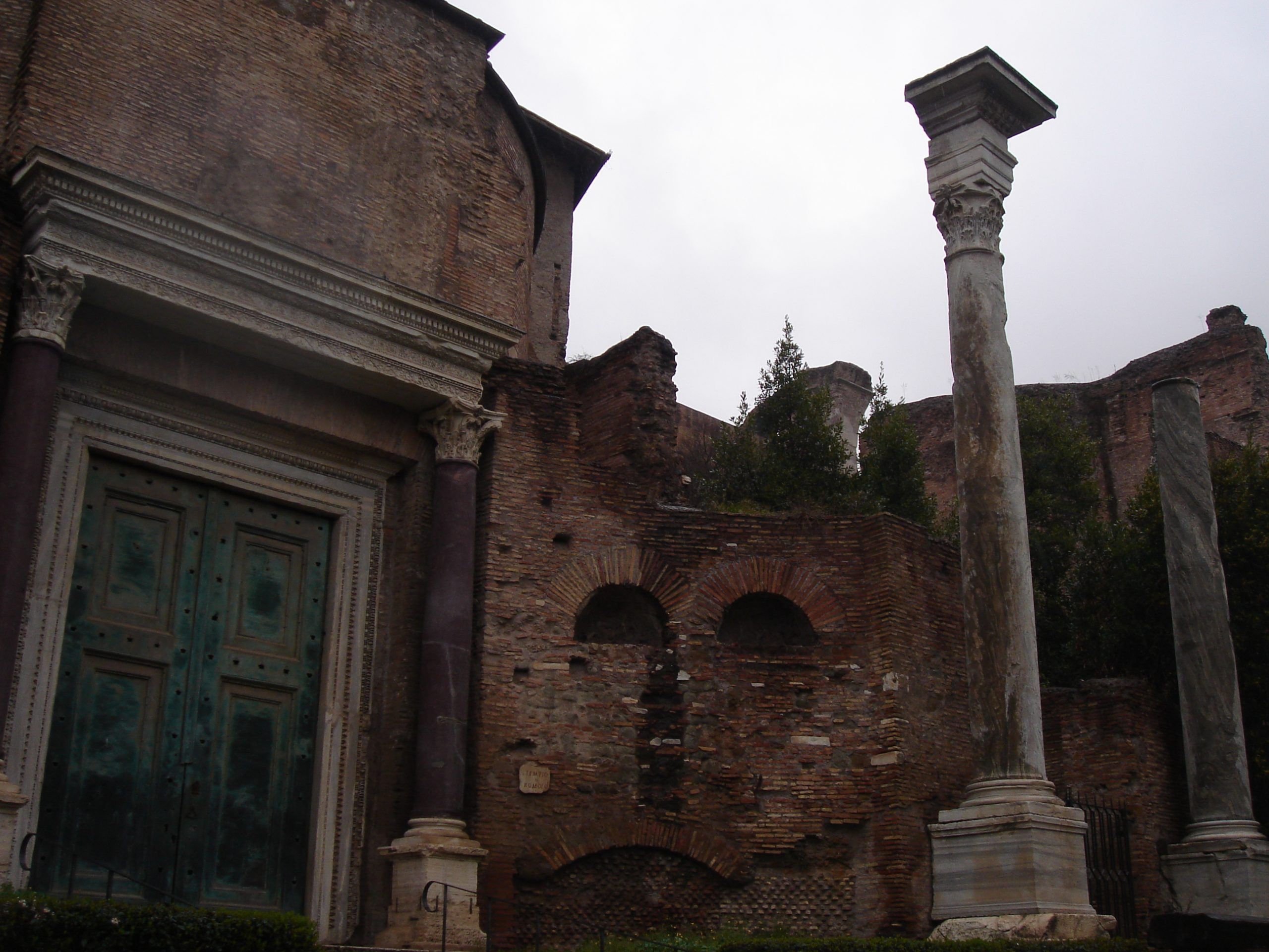 View of the Temple of Romulus in Rome, showing its green bronze doors, purple columns, and adjacent ruins with freestanding marble columns.