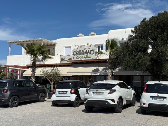 It's a low white building with a flat roof and rounded corners, typical of buildings in the Cyclades. It has an awning, and there are palm trees and bougainvillea out the front. Cars are parked in front of the building. Plastic signs on the left read TAVERNA and GRILL HOUSE in Comic Sans, and a more prominent central sign says OREGANO: Cook & Grill.