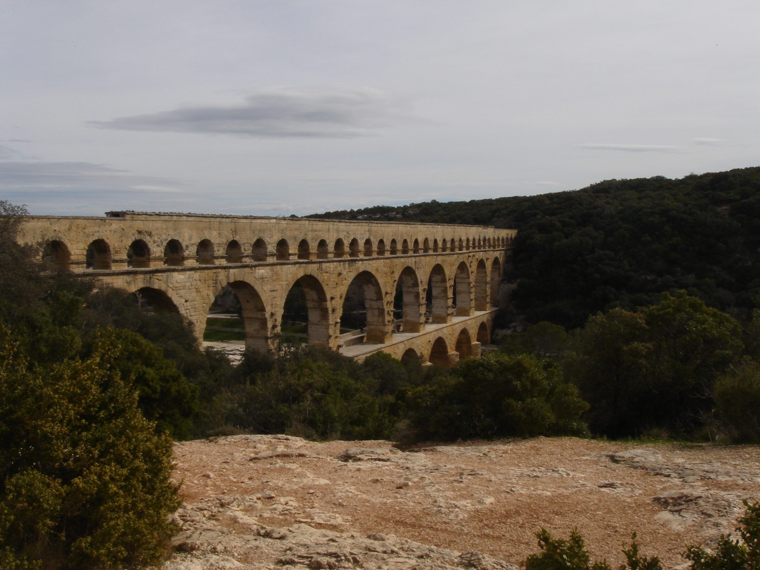 Wide view of the Pont du Gard aqueduct from a rocky plateau, with the structure framed by forested hills and cloudy sky.