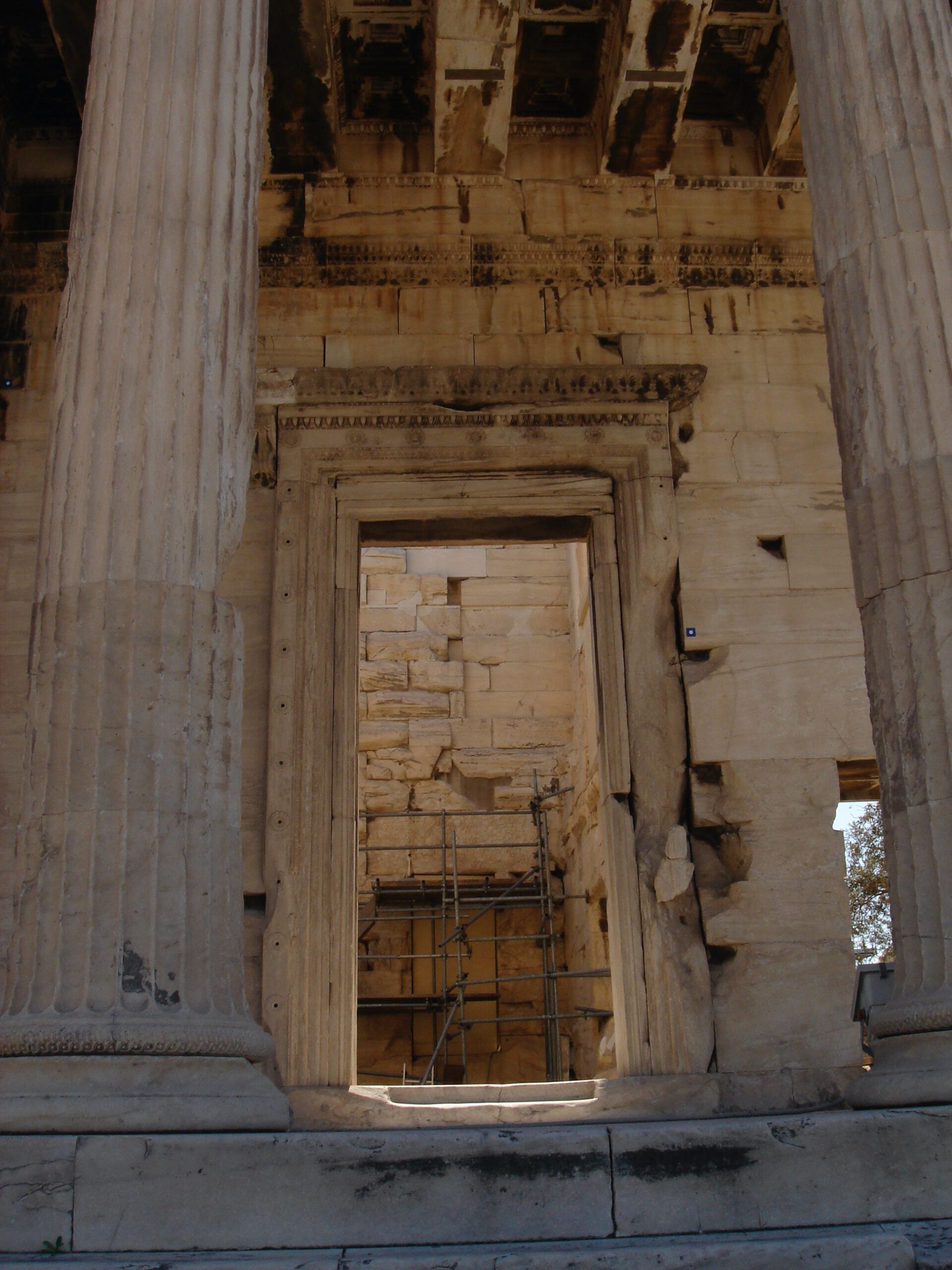 View through a doorway framed by tall fluted columns in the Propylaia of the Acropolis, showing interior scaffolding and weathered stone under the coffered ceiling.